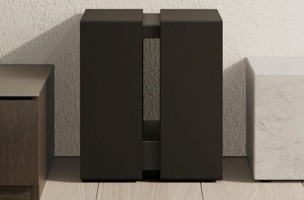 Czarny subwoofer o prostokątnym kształcie stoi na podłodze przy ścianie, otoczony z obu stron przez inne meble.