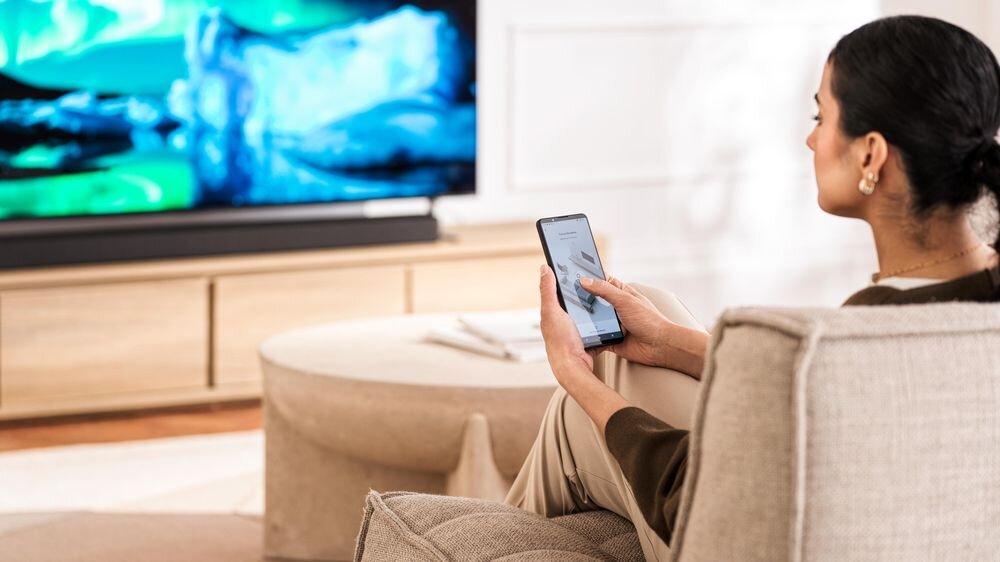 Osoba siedzi na sofie i trzyma w ręku smartfon, patrząc na włączony telewizor z kolorowym wyświetlaczem. W tle widoczny jest drewniany stolik i jasne wnętrze pokoju.