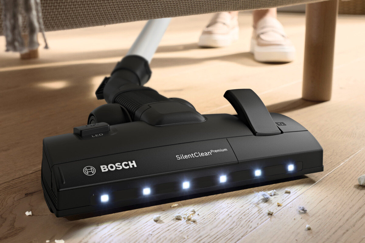 Czarna końcówka odkurzacza z logo BOSCH i napisem \'SilentClean Premium\' odkurza podłogę z rozsypanymi okruchami. Widoczne są diody LED na końcówce oraz fragment podłogi i mebla.