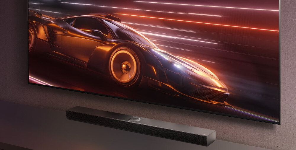 Na obrazie widoczny jest telewizor z dynamiczną grafiką przedstawiającą samochód wyścigowy oraz soundbar umieszczony poniżej ekranu. Soundbar jest czarny i ma prostokątny kształt.