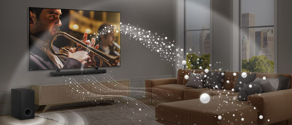 W salonie znajduje się duży telewizor z obrazem muzyka grającego na trąbce, a pod nim soundbar. Wokół soundbara i subwoofera widoczne są efekty wizualne symbolizujące dźwięk.