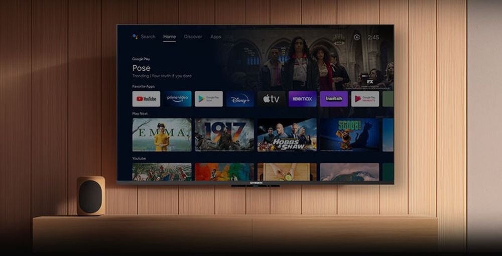 Telewizor wyświetla ekran główny z aplikacjami takimi jak YouTube, Prime Video, Disney+, Apple TV, HBO Max i innymi. Obok telewizora znajduje się głośnik na drewnianej półce.