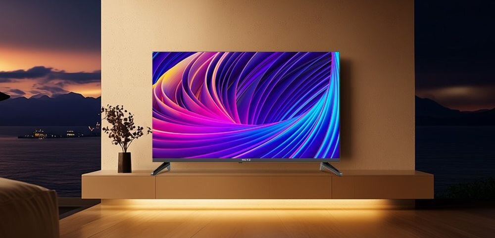 Telewizor z wyświetlaczem prezentującym kolorowy, abstrakcyjny wzór w odcieniach fioletu i niebieskiego, umieszczony na jasnej szafce. W tle widoczny jest krajobraz z zachodzącym słońcem.