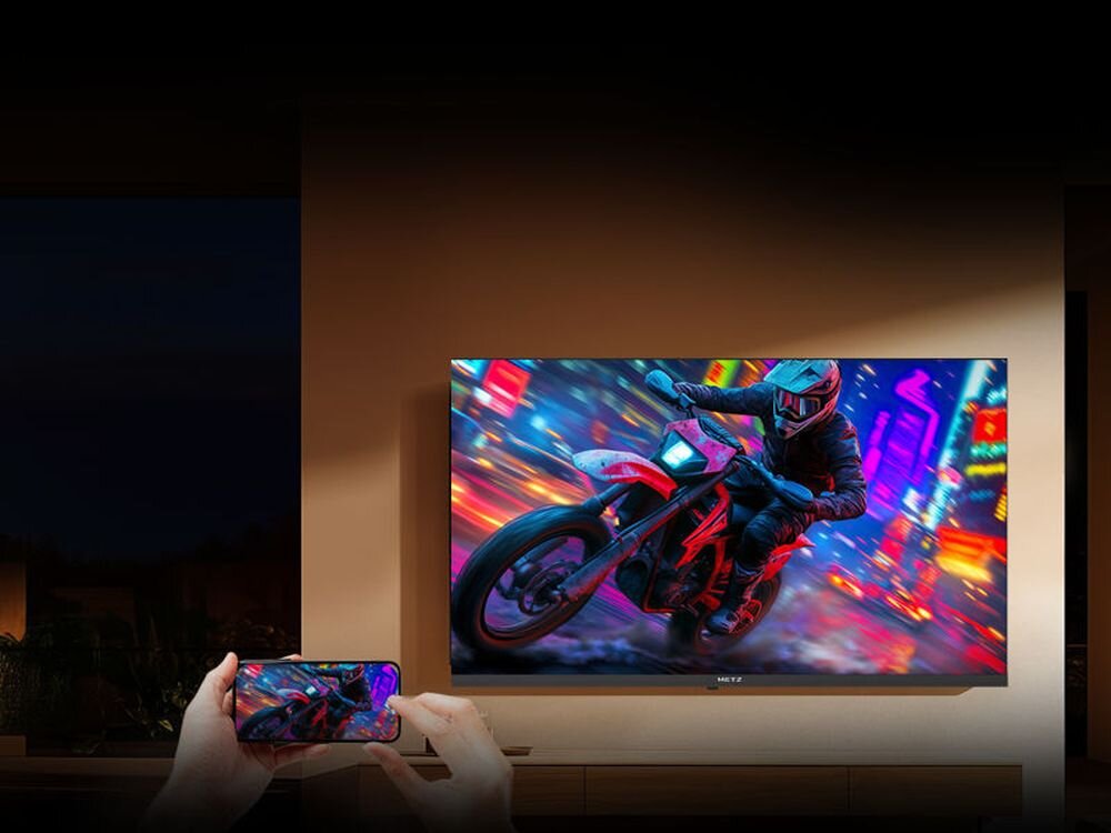 Na ścianie wisi telewizor wyświetlający dynamiczny obraz motocyklisty w kasku, jadącego przez kolorowe miasto. Przed telewizorem widoczne są ręce trzymające smartfon z tym samym obrazem na ekranie.