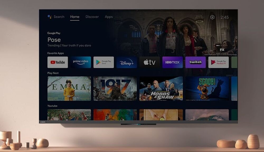 Telewizor wyświetla ekran główny z aplikacjami takimi jak YouTube, Prime Video, Disney+, Apple TV, HBO Max, Twitch i Google Play. Na półce pod telewizorem znajdują się dekoracyjne elementy.