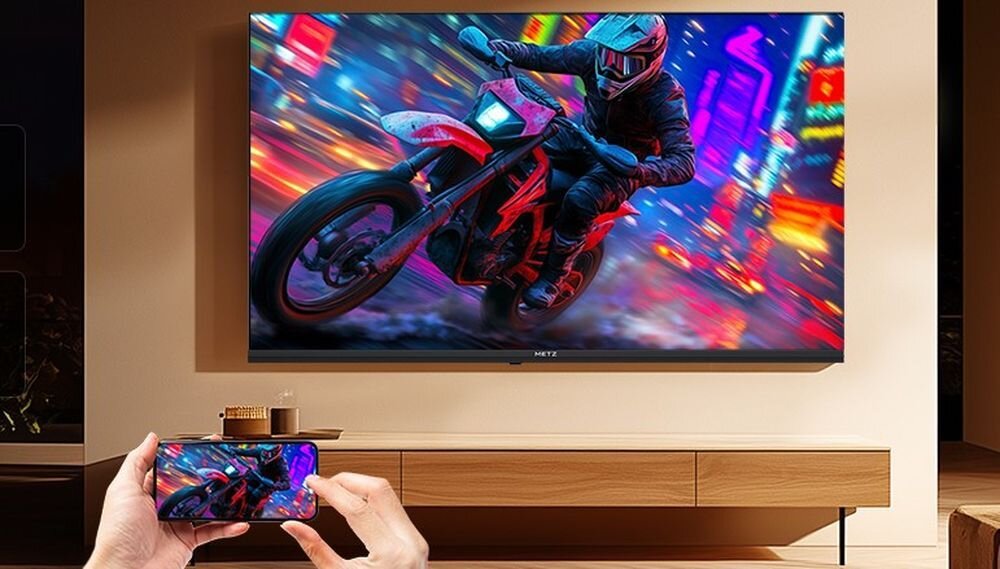 Na ścianie wisi duży telewizor wyświetlający dynamiczny obraz motocyklisty na tle kolorowego miasta. Przed telewizorem osoba trzyma smartfon, na którym widoczny jest ten sam obraz co na ekranie telewizora.