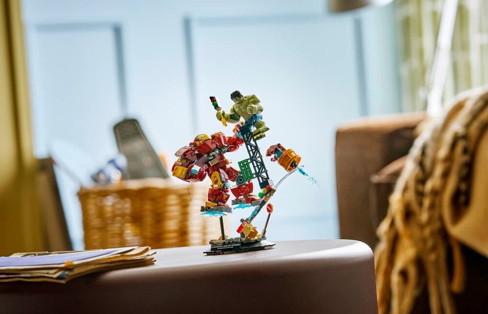 Na stoliku znajduje się zestaw LEGO przedstawiający scenę walki dwóch postaci: jednej w czerwonym pancerzu i drugiej zielonej. W tle widoczne są elementy wnętrza, takie jak kosz i kanapa.