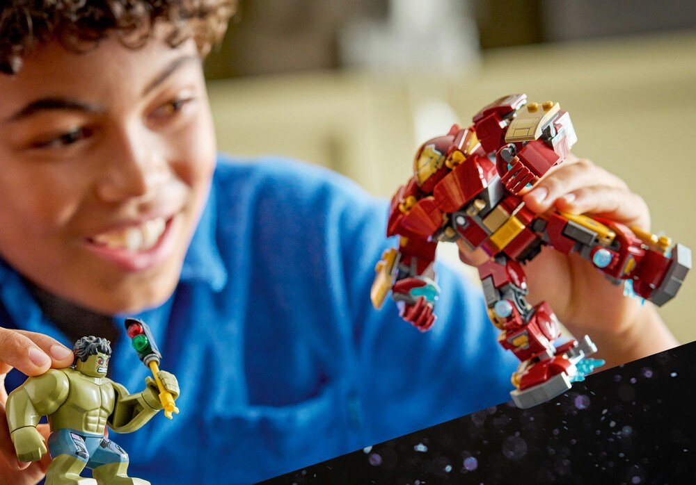 Dziecko bawi się figurkami LEGO przedstawiającymi postacie Hulk i Hulkbuster. Figurka Hulkbustera jest w kolorze czerwonym z żółtymi i szarymi elementami, a figurka Hulka jest zielona z fioletowymi spodenkami.