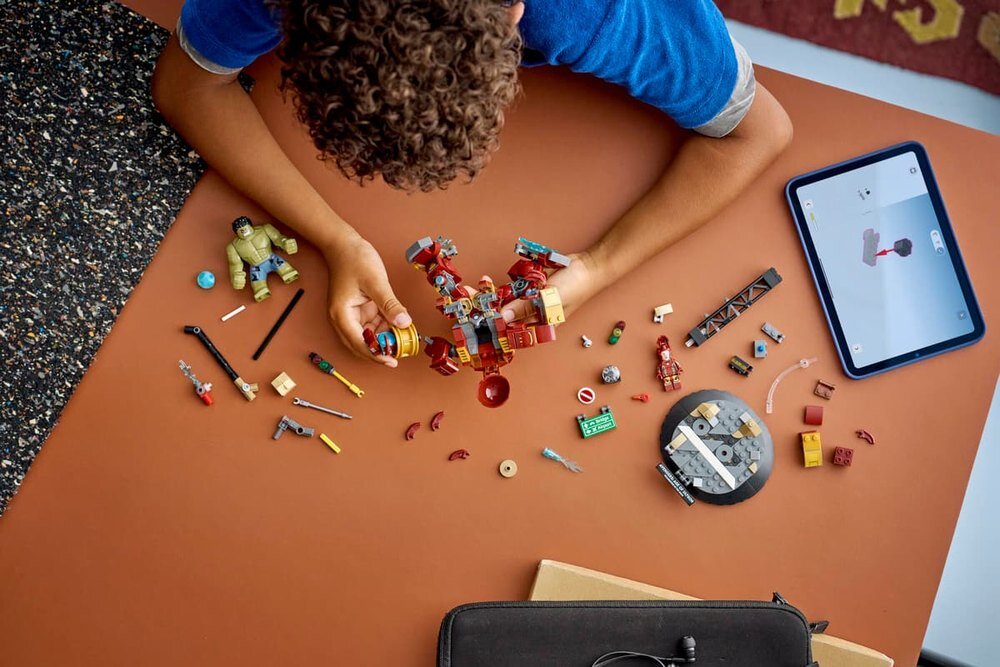 Dziecko składa zestaw klocków LEGO na brązowym stole, obok leży tablet z instrukcją. Na stole widoczne są różne elementy LEGO, w tym figurki i części konstrukcyjne.