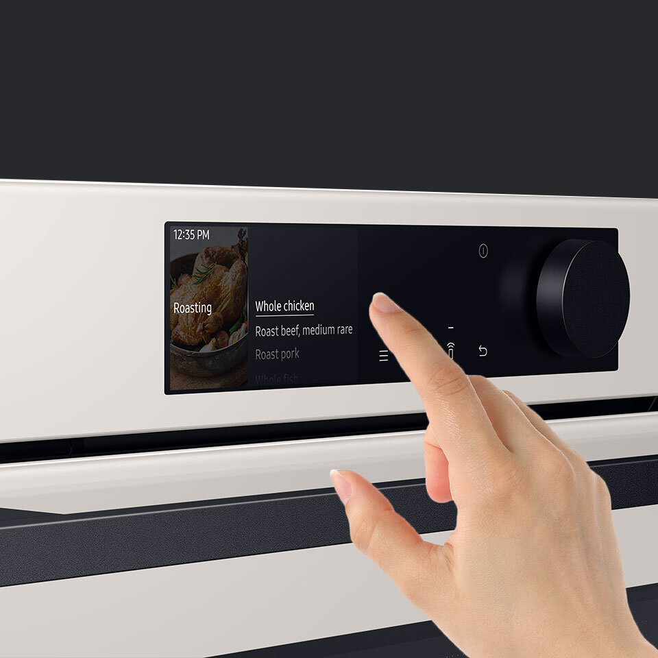 Na zdjęciu widać panel sterowania piekarnika z wyświetlaczem dotykowym, na którym widnieje tekst: \'12:35 PM\', \'Roasting\', \'Whole chicken\', \'Roast beef, medium rare\', \'Roast pork\'. Obok wyświetlacza znajduje się pokrętło, a ręka dotyka ekranu.