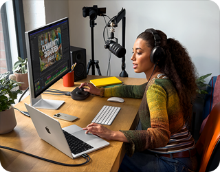 Osoba siedzi przy biurku z laptopem i monitorem, używając sprzętu audio. Na monitorze widoczny jest program do edycji wideo z napisem \'COMMUNITY GARDEN\'.