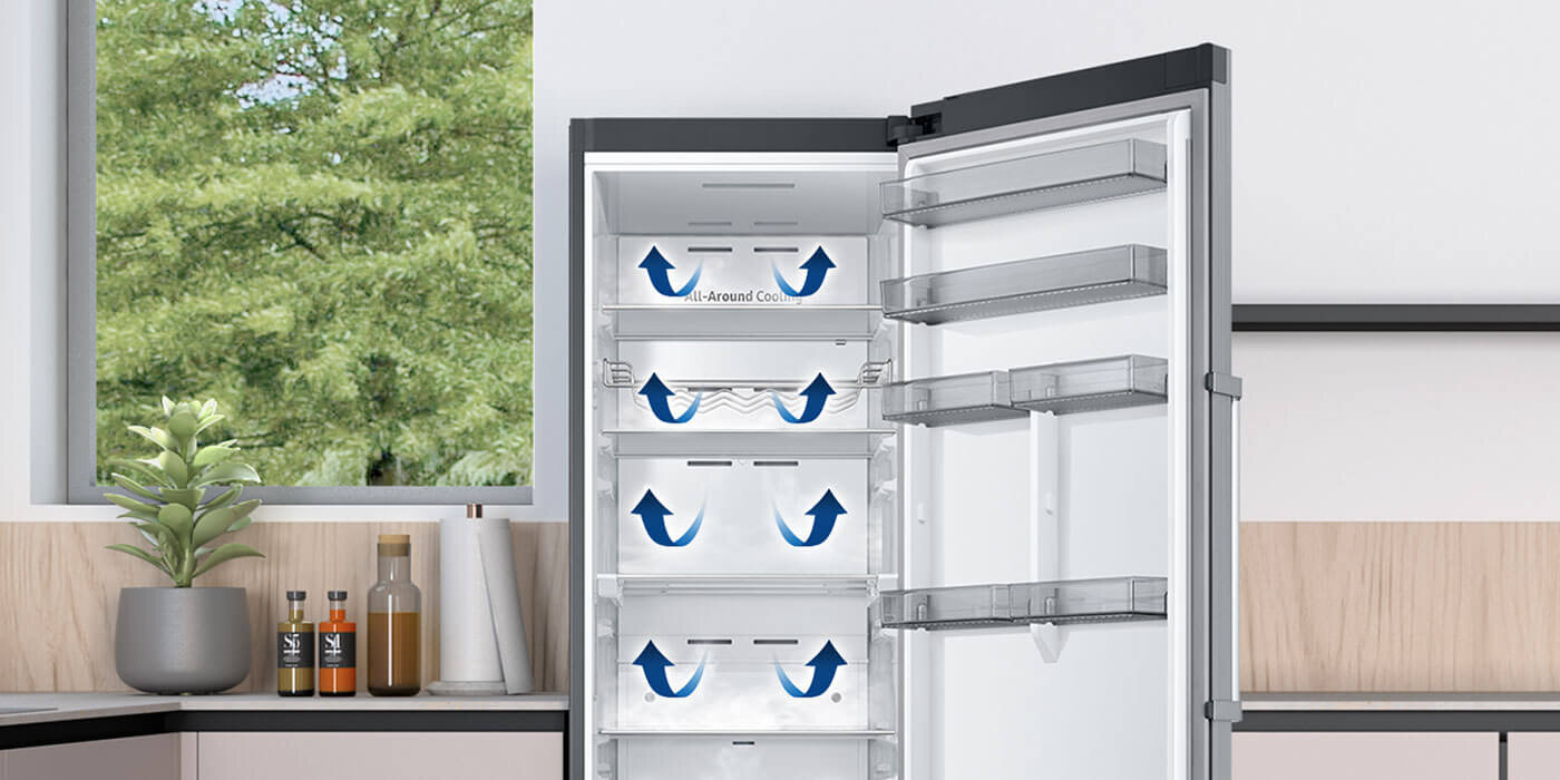 jednodrzwiowa chłodziarka z systemem All Around Cooling Otwarta lodówka z widocznymi półkami i strzałkami wskazującymi przepływ powietrza. W tle znajduje się okno z widokiem na drzewa oraz blat kuchenny z rośliną i butelkami.