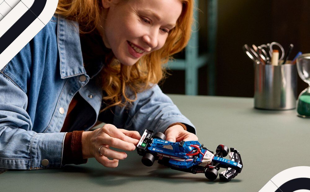 Osoba trzyma model bolidu F1 z klocków LEGO na stole. W tle widoczne są przybory biurowe w metalowym pojemniku.