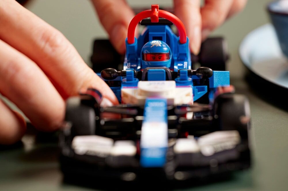 Na obrazie widoczny jest model samochodu wyścigowego z klocków LEGO w kolorach niebieskim, czarnym i czerwonym. W tle widać dłonie osoby, która trzyma model.