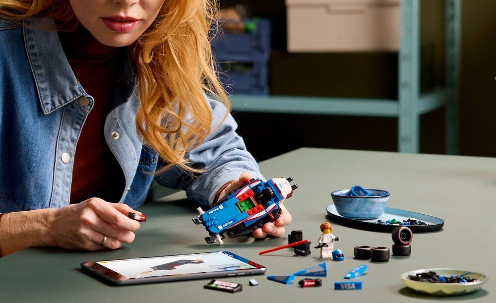 Osoba składa model samochodu z klocków LEGO na stole, obok leży tablet z instrukcją oraz różne elementy zestawu. Na stole widoczna jest karta z logo \'VISA\'.