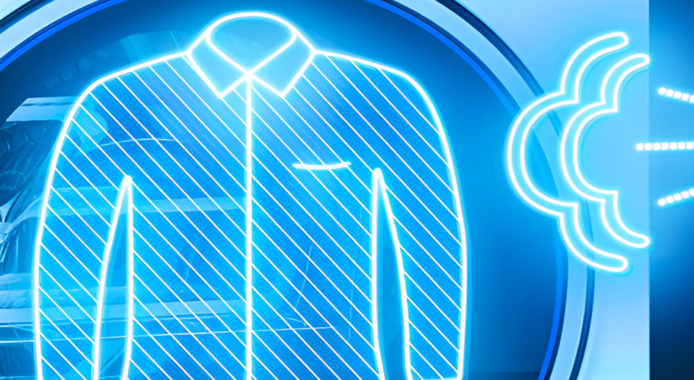 Grafika przedstawia niebieski, neonowy kontur koszuli z kołnierzykiem, obok której znajduje się symbol przypominający chmurę z promieniami. W tle widoczny jest fragment pralki.