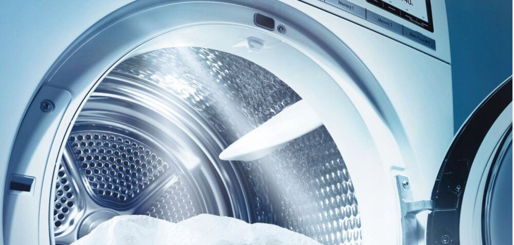 Obraz przedstawia wnętrze bębna pralki z otwartymi drzwiami, przez które wpada światło. Widać metalową strukturę bębna oraz fragment panelu sterowania z przyciskami i wyświetlaczem.