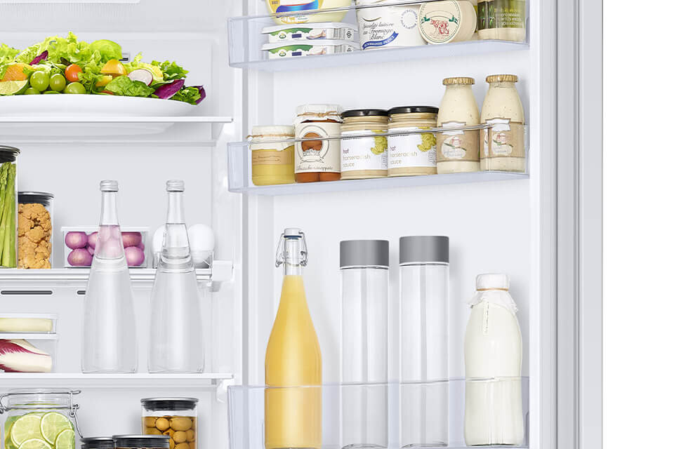 Dodatkowe miejsce na produkty na drzwiach lodówki Wnętrze lodówki z widocznymi półkami wypełnionymi różnymi produktami spożywczymi, takimi jak słoiki, butelki i pojemniki z jedzeniem. Na górnej półce znajduje się talerz z sałatką i owocami.