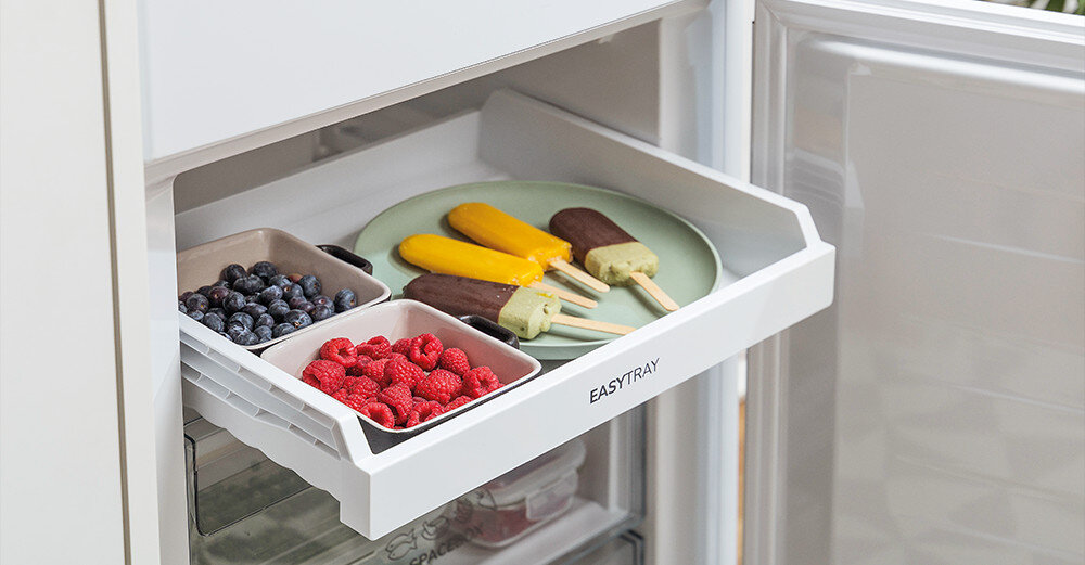 Otwarta lodówka z wysuniętą półką oznaczoną napisem \'EASYTRAY\', na której znajdują się owoce i lody na patyku. W tle widoczne są szuflady zamrażarki.