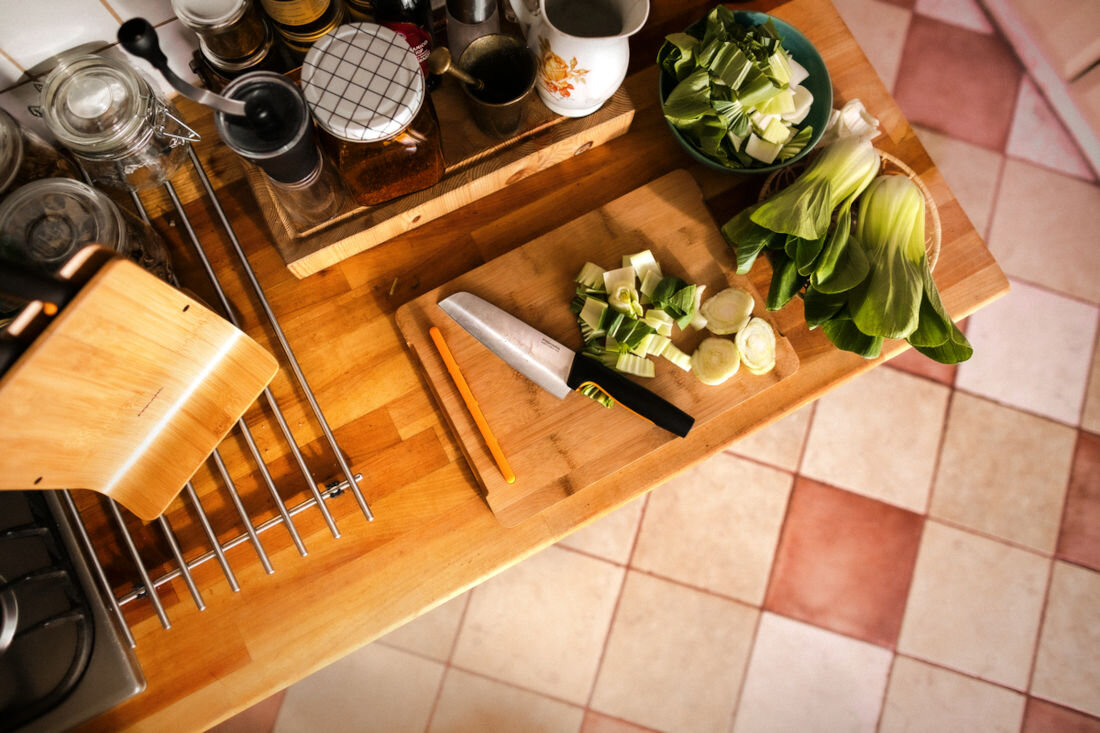 Na drewnianym blacie kuchennym znajduje się deska do krojenia z nożem i pokrojonymi warzywami. Obok leży świeża kapusta pak choi, a w tle widać różne pojemniki i akcesoria kuchenne.