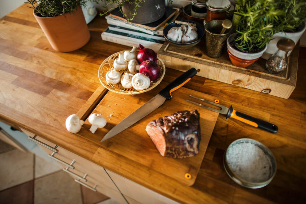 Na drewnianym blacie kuchennym znajduje się nóż i widelec do mięsa obok kawałka pieczeni. Obok leży talerz z pieczarkami i cebulą, a w tle widać doniczki z ziołami i przyprawy.