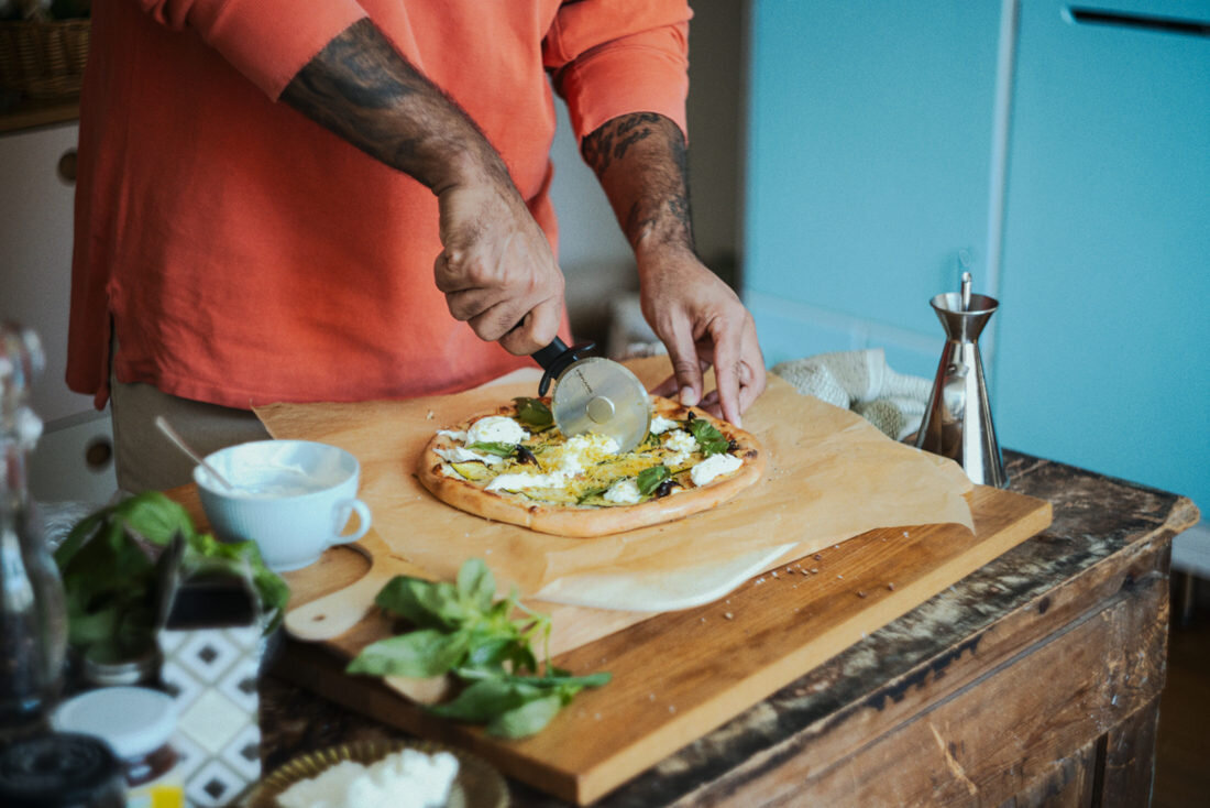 Osoba kroi pizzę nożem do pizzy na drewnianej desce. W tle widoczne są różne kuchenne akcesoria i niebieska lodówka.