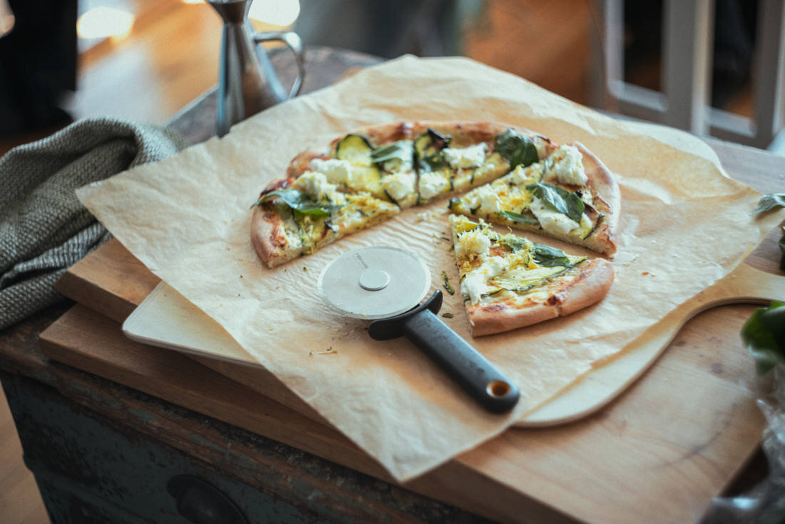 Na drewnianej desce leży pizza z zielonymi dodatkami i serem, obok znajduje się nóż do pizzy z okrągłym ostrzem. Pizza jest częściowo pokrojona i leży na papierze do pieczenia.