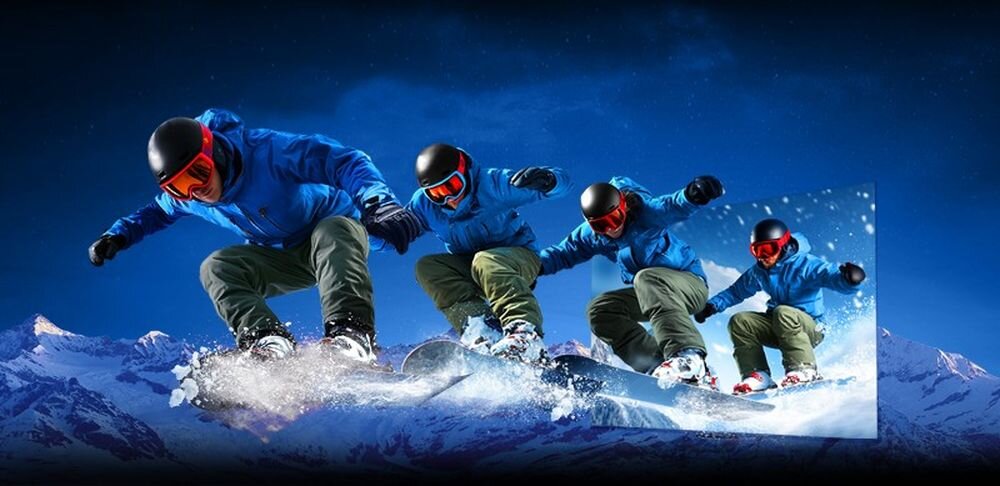 Czterech snowboardzistów w niebieskich kurtkach i czerwonych goglach zjeżdża po śniegu na tle górskiego krajobrazu. Obraz jest dynamiczny, z efektem rozmycia śniegu.