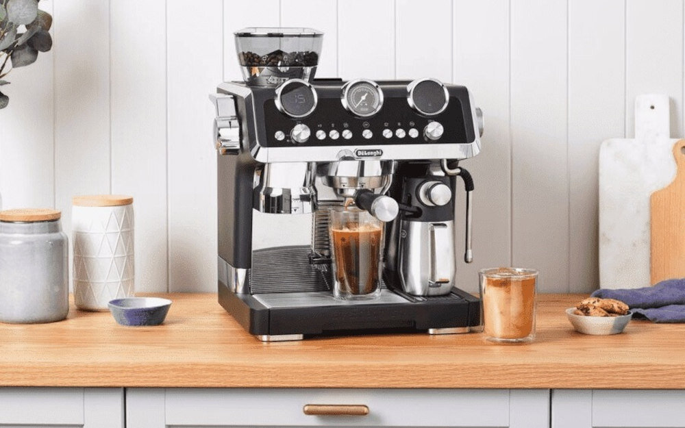 Na drewnianym blacie stoi ekspres do kawy z logo \'DeLonghi\', obok którego znajdują się dwie szklanki z kawą. W tle widoczne są różne akcesoria kuchenne, takie jak słoiki i deski do krojenia.