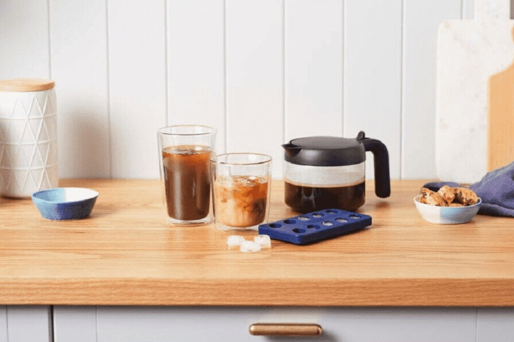 Na drewnianym blacie stoją dwie szklanki z napojami, dzbanek z czarnym uchwytem, niebieska tacka na kostki lodu oraz miseczki z przekąskami. W tle widoczna jest biała ściana z pionowymi panelami.