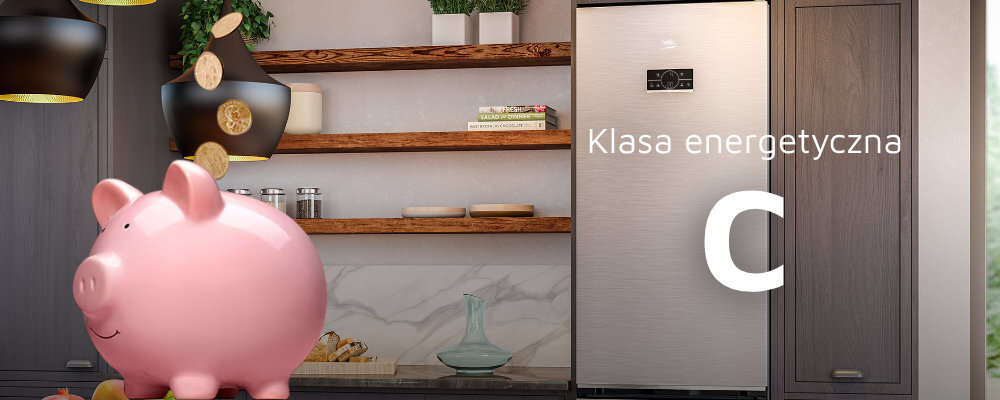 W kuchni znajduje się lodówka w kolorze ciemnego inoxu, obok której widnieje napis \'Klasa energetyczna C\'. Na pierwszym planie stoi różowa skarbonka w kształcie świnki, a w tle widać drewniane półki z dekoracjami.