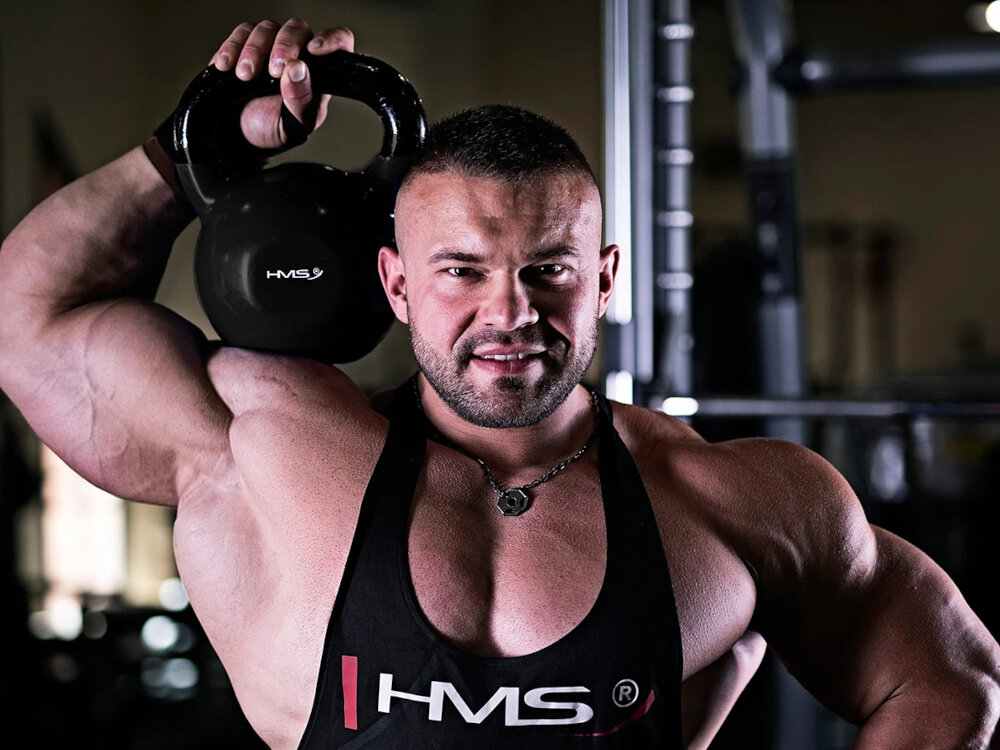 Kettlebell HMS KNV32 (32 kg) siłownia umięśniona osoba trzyma kettlebell na ramieniu w tle sprzęty do ćwiczeń trening siłowy trening kondycyjny wszechstronne zastosowanie zdrowie