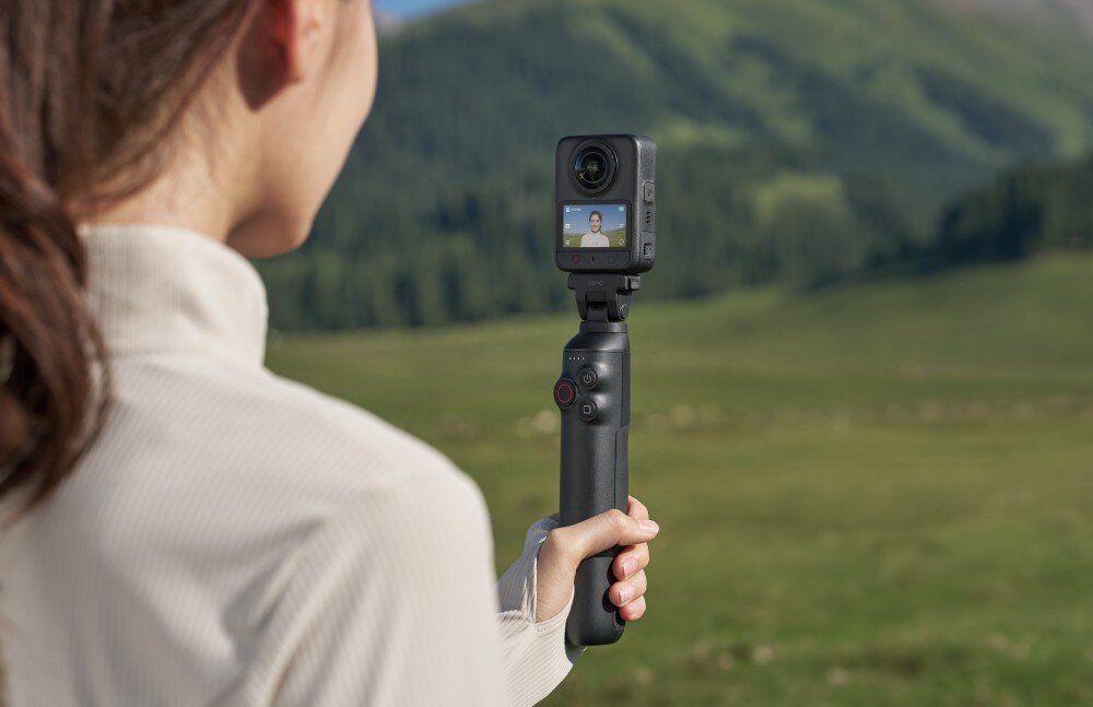 Wysięgnik z wbudowanym akumulatorem DJI CP.OS.00000637.01 Osmo 360 II Kobieta trzyma w dłoni uchwyt z zamontowaną małą kamerą sferyczną na tle zielonego wzgórza, na ekranie urządzenia widać jej twarz