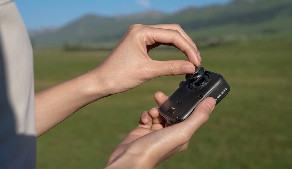 Kamera sportowa DJI Osmo 360 II Dłonie nakładające lub zdejmujące okrągłą osłonę obiektywu na małe urządzenie na tle zielonej przestrzeni