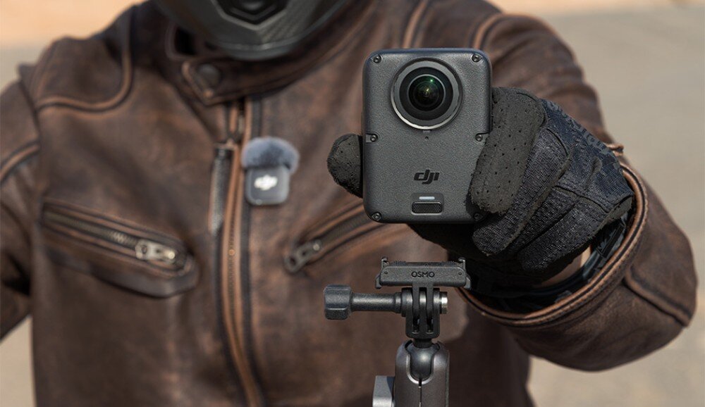 Kamera sportowa DJI Osmo 360 II Osoba w skórzanej kurtce i rękawicy trzyma małe urządzenie nagrywające zamontowane na uchwycie motocyklowym