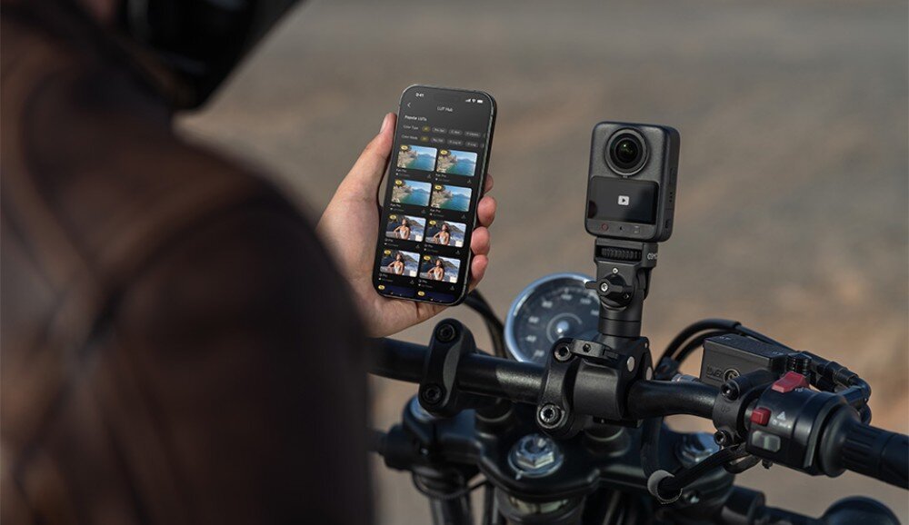 Kamera sportowa DJI Osmo 360 II Dłoń trzymająca smartfon z otwartą aplikacją do edycji wideo obok małego urządzenia zamontowanego na kierownicy motocykla
