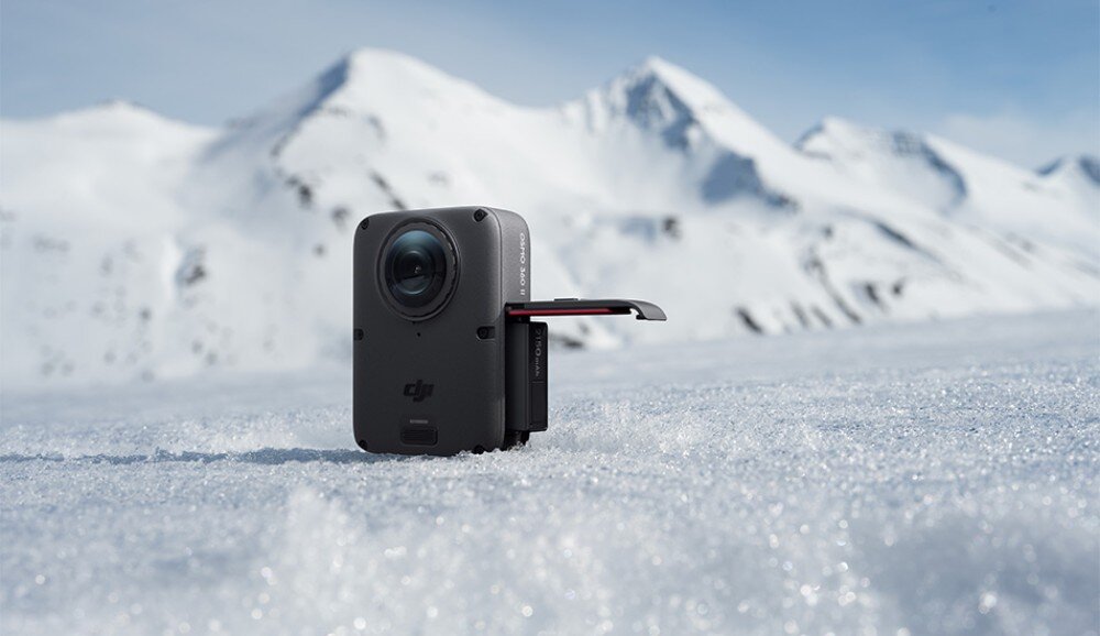Kamera sportowa DJI Osmo 360 II Małe urządzenie stojące na ośnieżonym podłożu z otwartą komorą i wysuniętym akumulatorem, w tle ośnieżone szczyty górskie