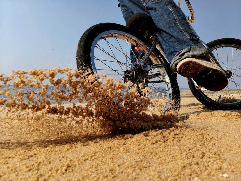 Osoba na rowerze jedzie po piaszczystej plaży, wzbijając piasek w powietrze. W tle widać jasne niebo.