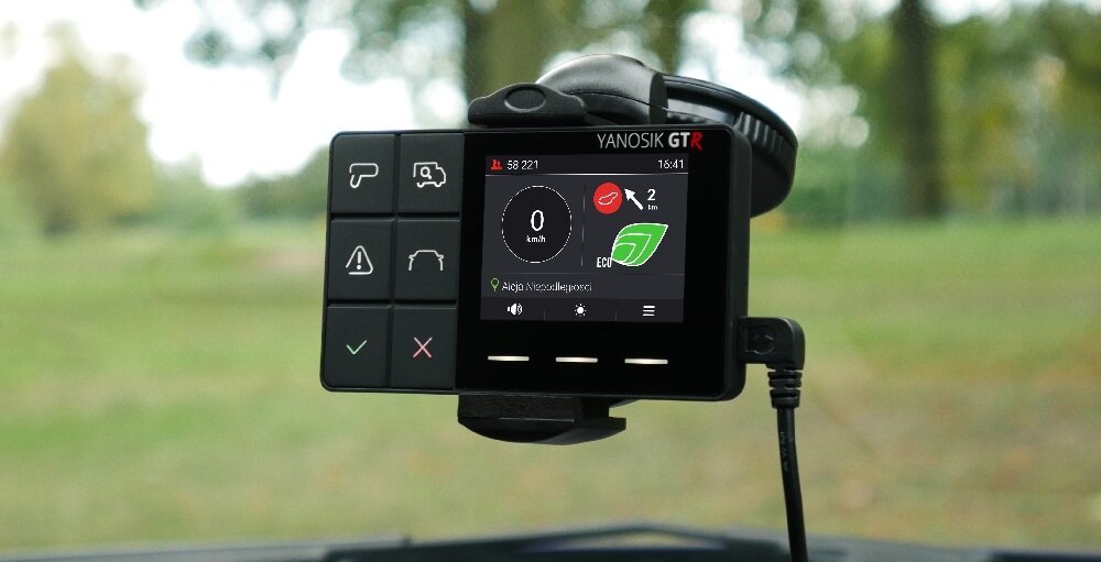 Urządzenie elektroniczne z ekranem wyświetlającym prędkość 0 km/h oraz ikonę zielonego liścia, zamontowane na desce rozdzielczej samochodu. Na obudowie widoczne są przyciski z symbolami oraz napis \'YANOSIK GTR\'.