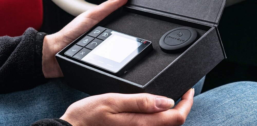 Osoba trzyma otwarte pudełko z urządzeniem elektronicznym z wyświetlaczem i kilkoma przyciskami oraz okrągłym elementem z napisem \'YANBOX\'.