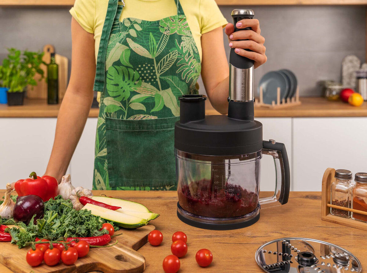 Osoba w zielonym fartuchu obsługuje blender stojący na blacie kuchennym, w tle widoczne są naczynia i przyprawy. Na blacie znajdują się różne warzywa, w tym pomidory, papryka i cukinia.