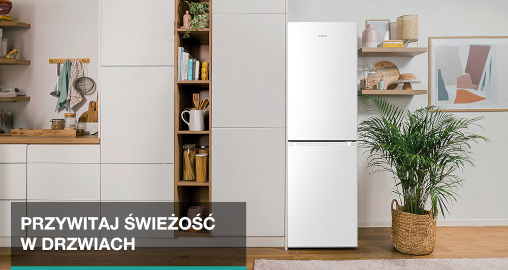 Biała lodówka GORENJE stoi w nowoczesnej kuchni obok rośliny w wiklinowym koszu. Na dole obrazu widnieje tekst: \'PRZYWITAJ ŚWIEŻOŚĆ W DRZWIACH\'.
