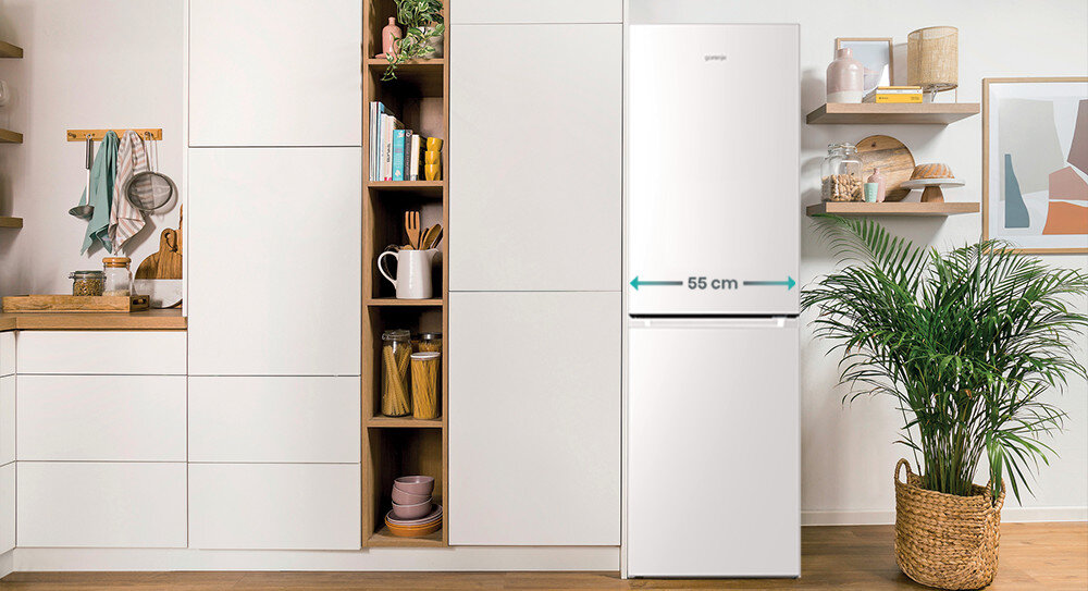 Biała lodówka z napisem \'gorenje\' stoi w nowoczesnej kuchni obok regału z książkami i naczyniami. Na lodówce widoczny jest napis \'55 cm\'.