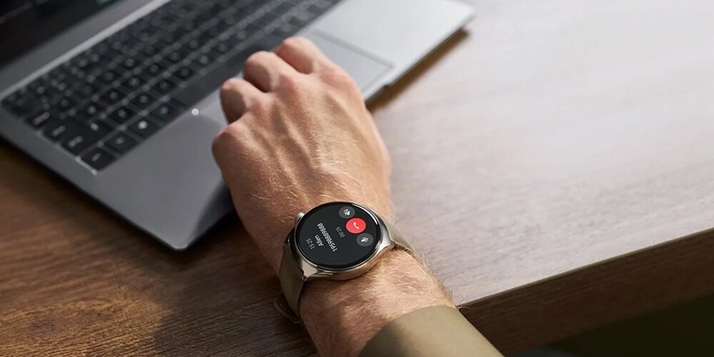 Osoba nosi smartwatch na nadgarstku, który wyświetla ekran z informacjami o połączeniu przychodzącym. W tle widoczna jest klawiatura laptopa na drewnianym biurku.