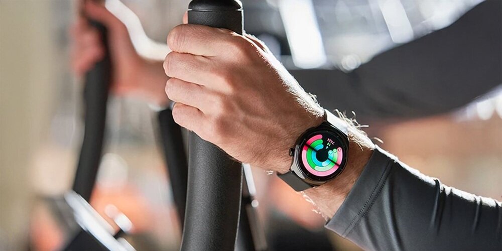 Osoba trzyma uchwyt urządzenia do ćwiczeń, na nadgarstku ma smartwatch z kolorowym wyświetlaczem pokazującym okrągłe wskaźniki aktywności.