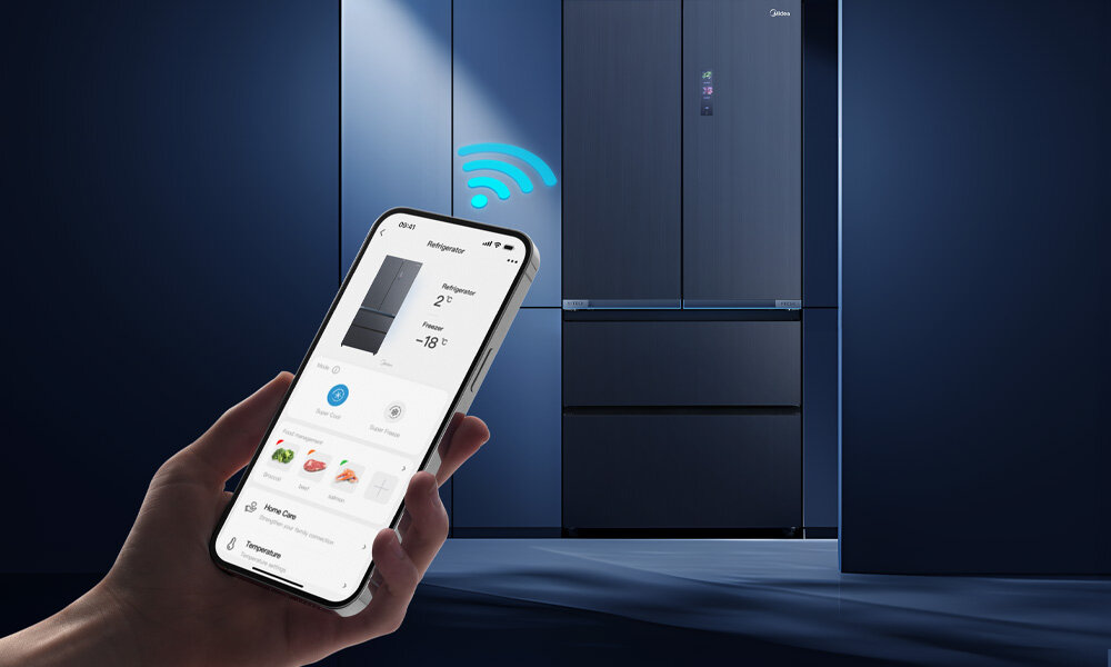 Osoba trzyma smartfon z aplikacją pokazującą temperaturę lodówki, w tle widoczna jest szara lodówka z logo Midea i ikoną sygnału Wi-Fi.