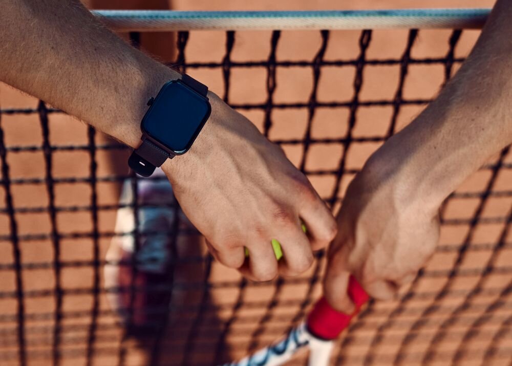 Osoba nosi czarny smartwatch na nadgarstku, trzymając rakietę tenisową i piłkę przy siatce na korcie tenisowym.