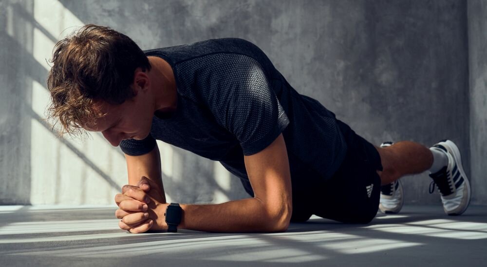 Osoba w sportowym stroju wykonuje ćwiczenie w pozycji deski, na nadgarstku ma czarny smartwatch. W tle widać szarą ścianę i padające cienie.