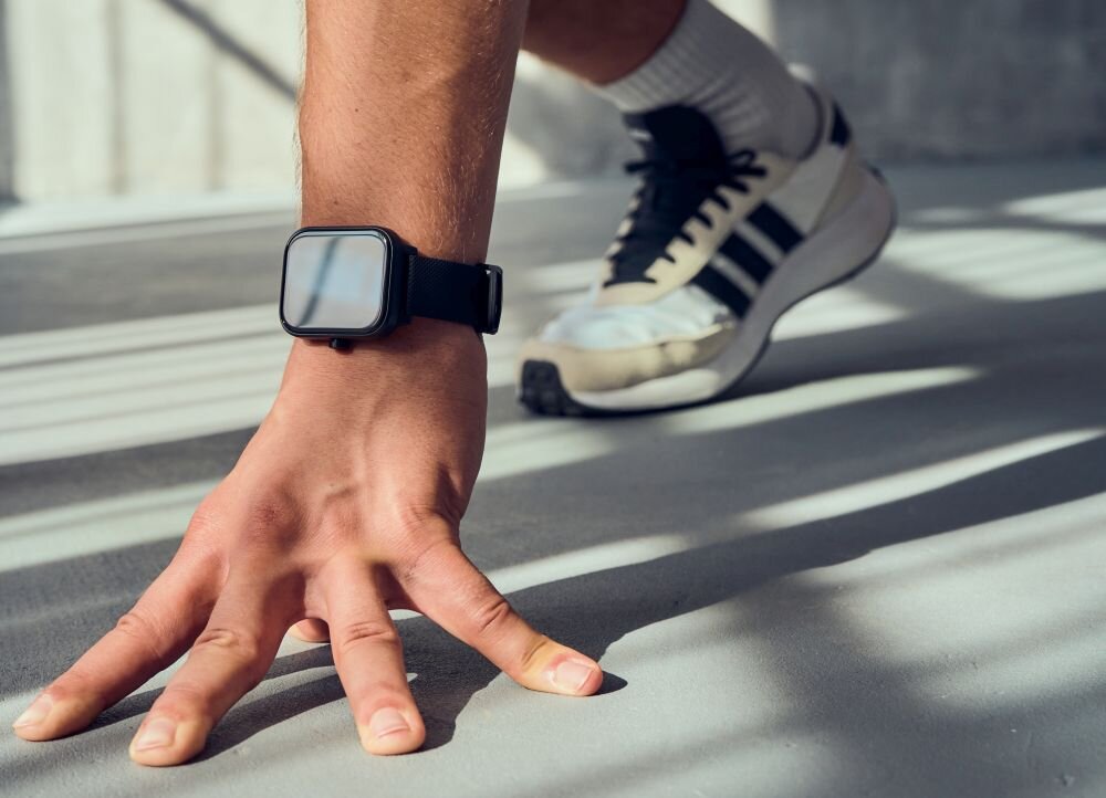 Osoba w sportowym obuwiu wykonuje ćwiczenie, mając na nadgarstku czarny smartwatch z prostokątnym ekranem.