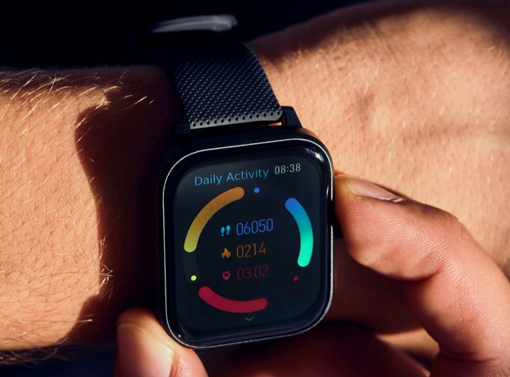 Na zdjęciu widoczny jest smartwatch z czarnym paskiem na nadgarstku, wyświetlający ekran z informacjami o aktywności dziennej: 06050 kroków, 0214 kalorii, 03.02 km. Na ekranie widnieje również napis \'Daily Activity\' oraz godzina 08:38.